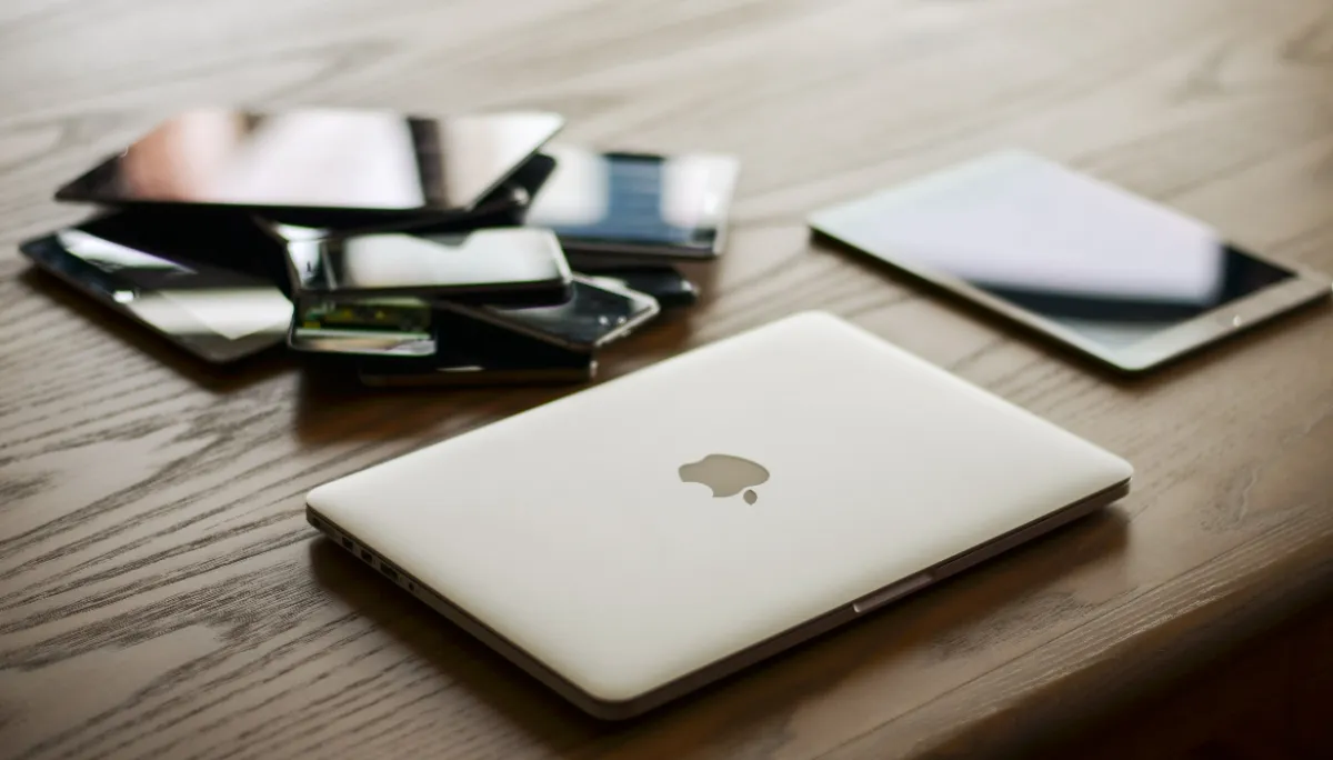 A closed laptop on a wooden desk beside a stack of tablets and phones